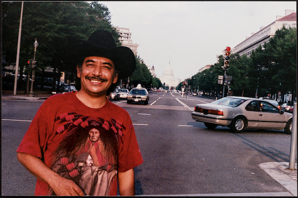 A smiling man with dark hair and mustache, black cowboy hat, red t-shirt illustrated with two Indigenous figures, faces the camera on the left side of the frame. In the background, a long avenue at the end of which is the U.S. Capitol.