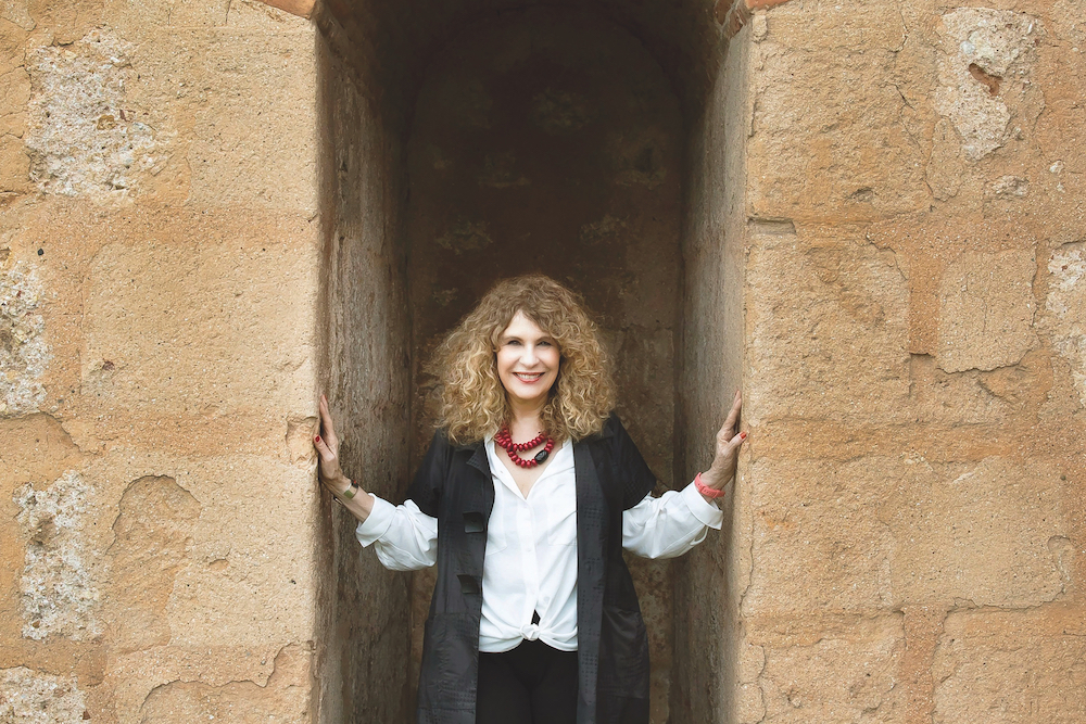 A woman in her sixties with light brown wavy hair stands at a stone wall at a place where the wall separates. Her hands rest on the parts of the wall beside her on each side. She smiles at the camera. She is wearing a white long-sleeved shirt, black short-sleeved jacket and pants, and a bright red necklace made of two strands of large beads.