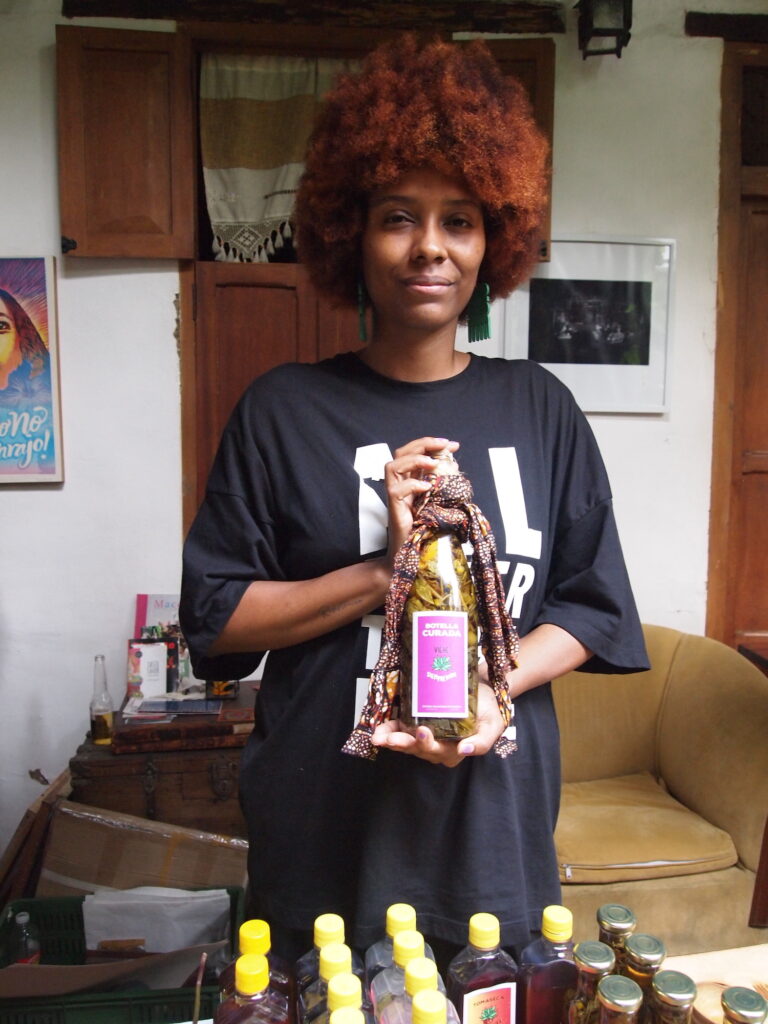 A Black woman holds a bottle and faces the camera. The bottle contains greenish-yellow herbs and liquid and has a pink label. There is a batiked cloth tied around the top. The woman's earrings are large turquoise hair combs and her hair is natural, very full, and reddish.