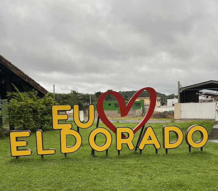 Large yellow letters placed on a green lawn spell out I Love Eldorado (in Portuguese). There is a fanciful outline of a bright red heart in the design. These letters are on a green lawn. The sky is wide an gray and cloudy in the background and above.