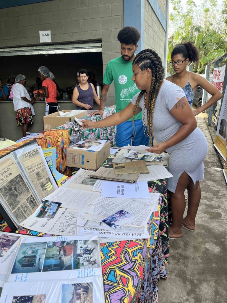 A table display shows old newspaper clippings, photo albums, notebooks, and papers. A Black woman reaches toward the table, placing items. Two other young Black people—a man and woman—stand near her. In the background, there is a counter labeled "Bar" where people in hair nets appear to be setting up food.