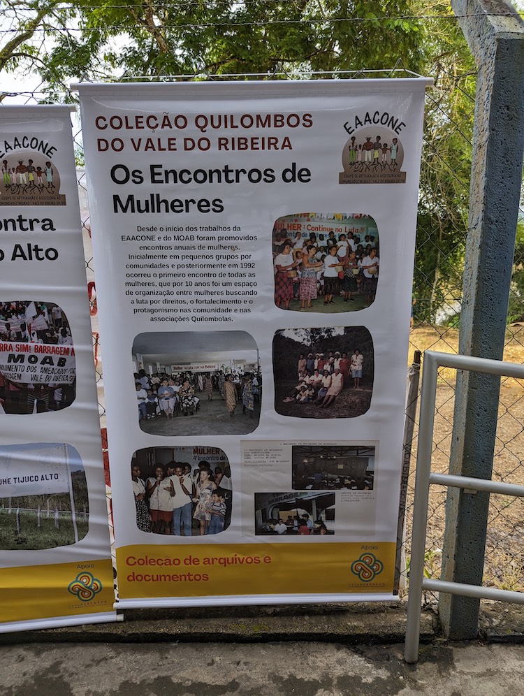 A large white vinyl sheet hangs from a chainlink fence. It is printed with information in Portuguese and photos from the EAACONE archive named Quilombos of Vale do Ribeira Collection. The photos and text are related to women's meetings. LLILAS Benson is cited at the bottom of the vinyl sheet as a sponsor.