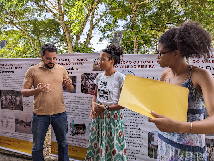 Three people stand in front of a chain link fence hung with vinyl posters, part of the traveling exhibition describing the EAACONE archive. On the left is a man, who is talking, in the middle, a woman whose t-shirt reads "Fight Like a Black Woman" (in Portuguese) and on the far right, another woman holds a yellow folder and is looking toward the man.