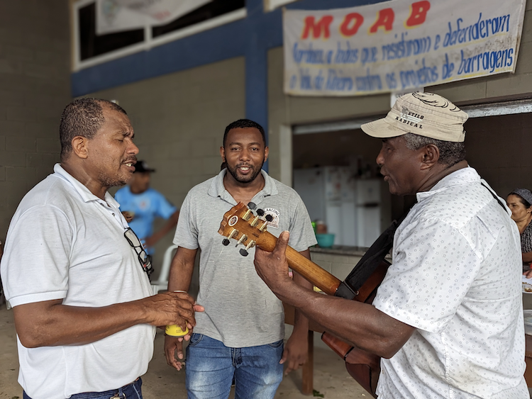 Three middle-aged Black men stand in a circle singing. The man on the right is playing a guitar. In the background, a banner hanging on the wall talks about MOAB, the historic anti-dam movement that has been a source of activism among quilombola communities in Vale do Ribeira.