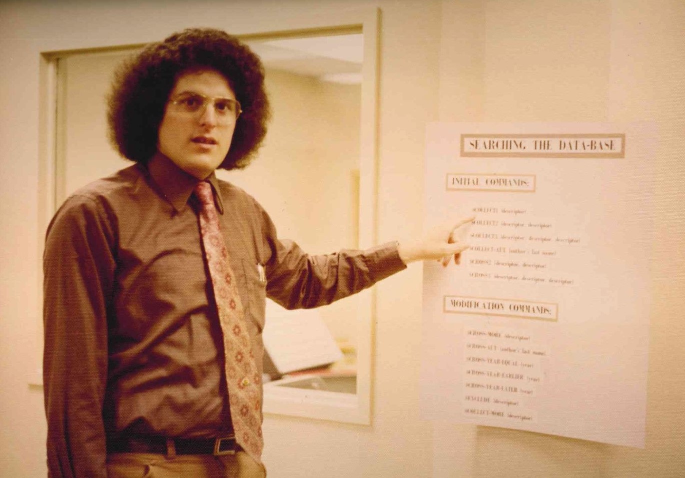 Library staff member gestures to a poster titled "Searching the Database" that lists database queries.