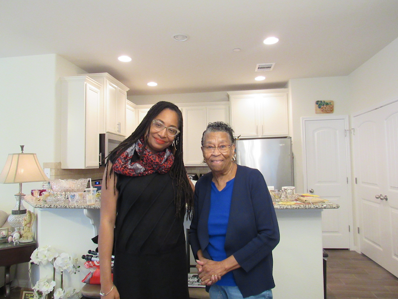 Two women pose together in a home, with a kitchen in the background. On the left is a younger Black woman with long dreads, glasses, and red lipstick who is wearing a red-white-and-black scarf; on the right an older Black woman in a bright blue top and dark blue blazer. Both are smiling.