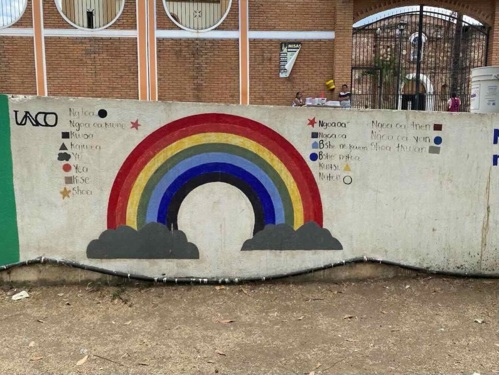 A pale-gray wall is in the foreground. Painted on the wall is a simple rainbow with dark-gray clouds at each end. On either side of the rainbow, small blocks of different colors are painted, each with a word next to it in black lettering. In the background there is a red brick building with black wrought-iron gates in an archway. A few people are also visible in the background.