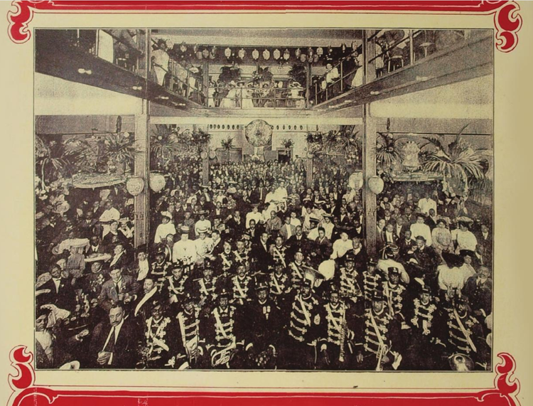 Photograph of the inside of Pekin Theater from 1905. There is decorative red border around an image of a large room with a balcony. There's a marching band in the foreground and hundreds of spectators. Everyone is facing the camera. 