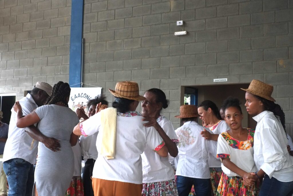 Black women of various ages (plus one man) dance in pairs. The women are almost all wearing white blouses. Some wear straw hats. Some wear colorful flowered skirts.