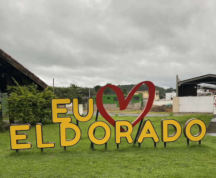 Grandes letras amarelas instaladas numa grama verde falam "Eu Amo Eldorado." Em vez da palavra "amo" tem um coração vermelho.