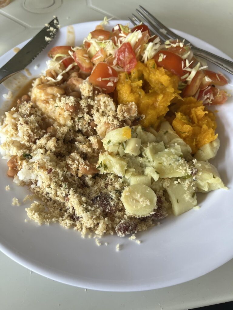A white plate piled high with colorful and tasty-looking food, including rice and beans topped with farofa, salad with tomatoes, orange pumpkin, and pale yellow yucca. A knife and fork rest on the edge of the plate.