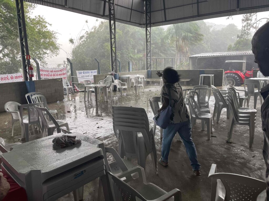 A quilombola elder runs, her back to the camera, arm oustretched, on a covered patio. Rain and wind are strong beyond the patio's cover, and the patio floor is wet. The wind blows her hair.